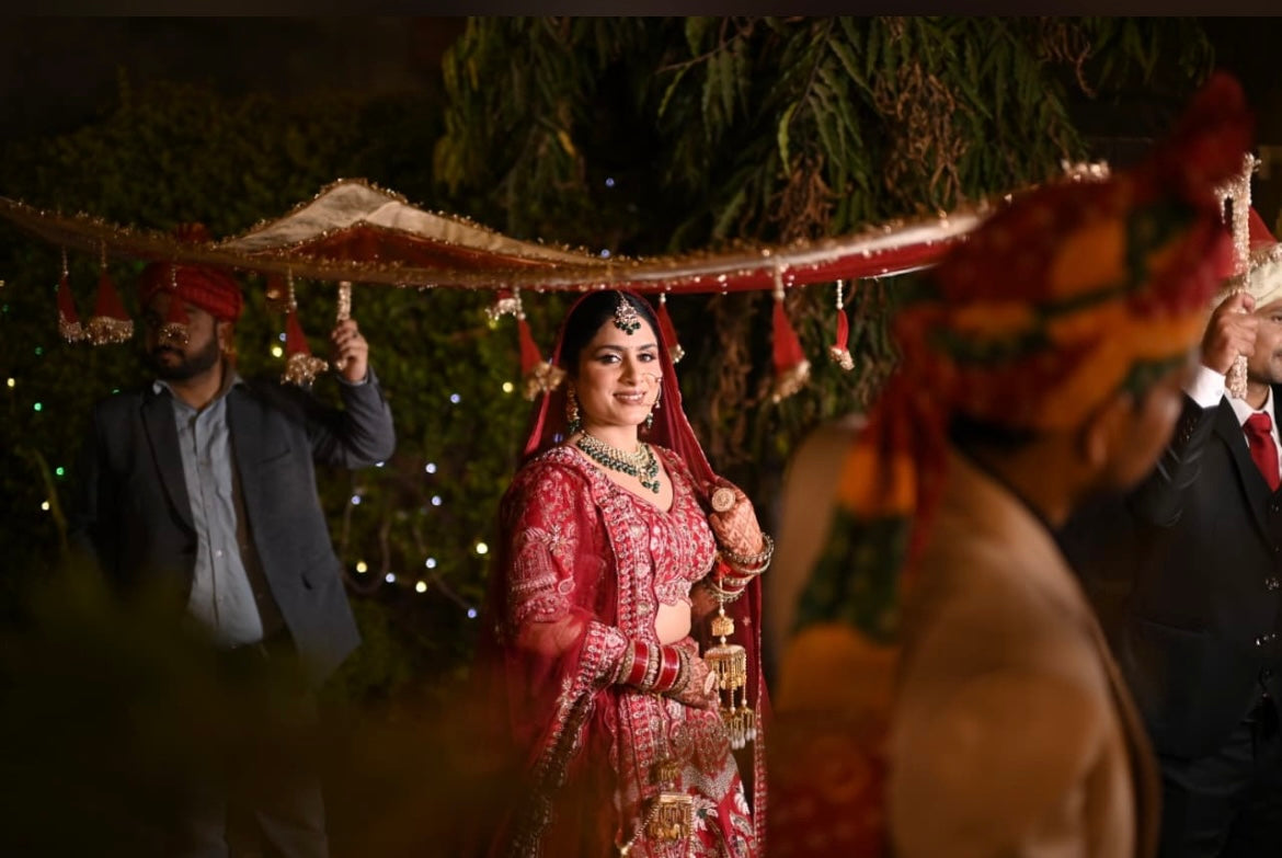 Woman in traditional red bridal outfit with jewelry, surrounded by people in formal attire, against a dark background.
